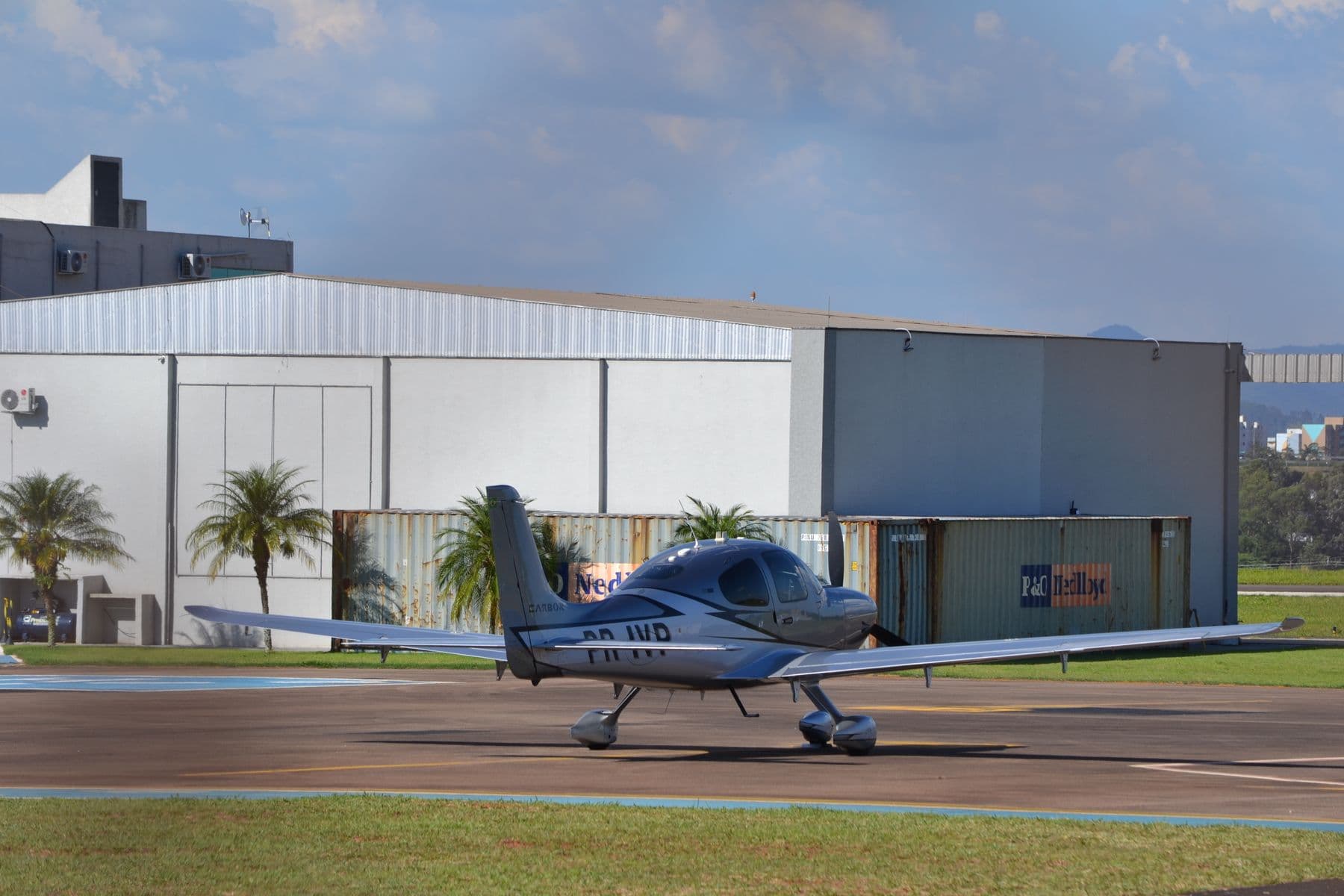 A small single-engine plane on the apron in front of a metal hangar, with palm trees behind.