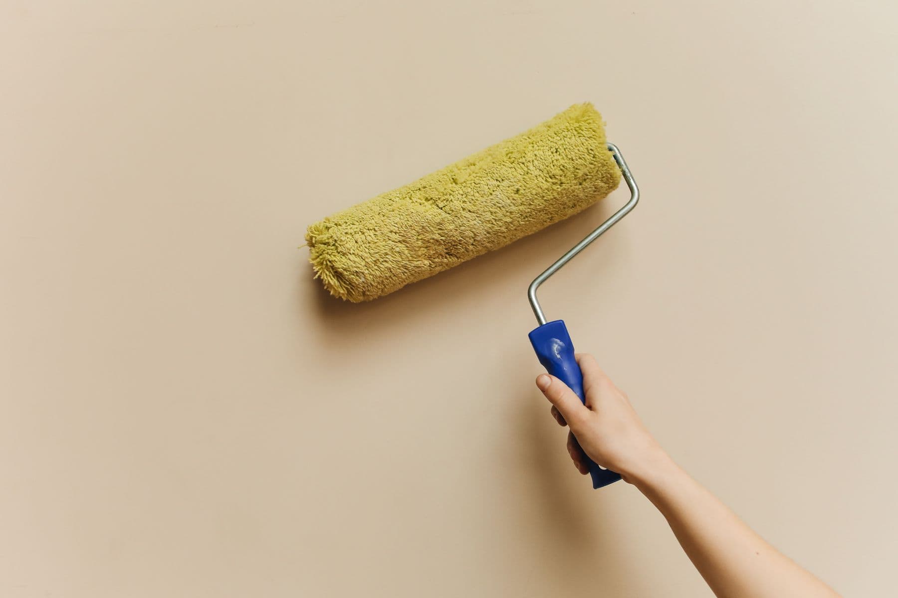 A hand rolling a yellow paint roller on a beige wall.