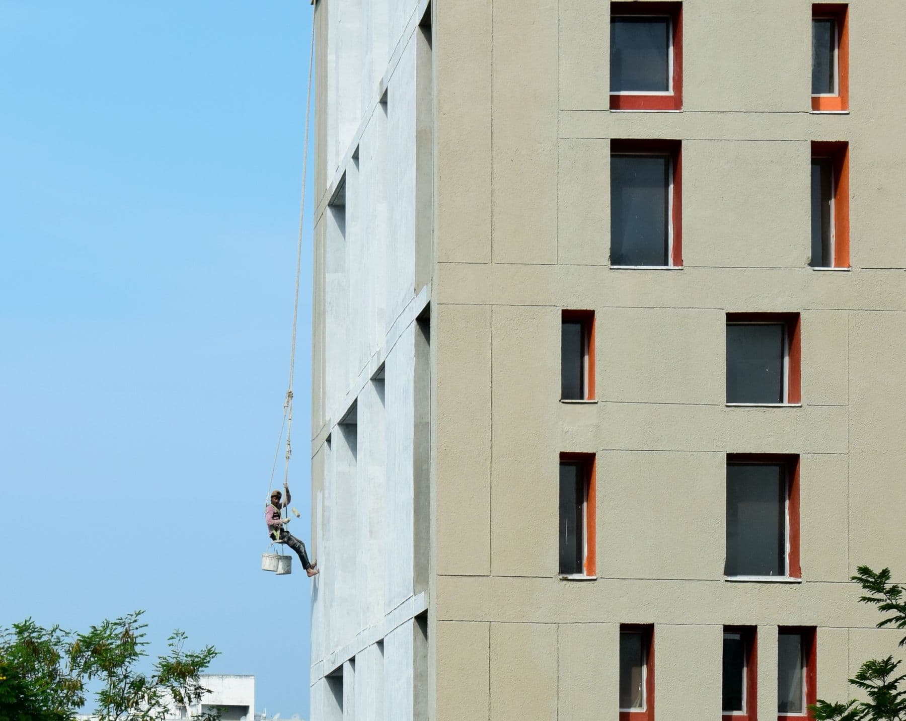 A painter on a rope seat hanging beside the tan wall of a tall apartment building.