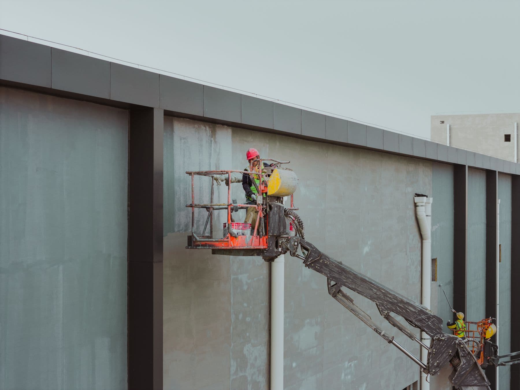 A worker in a hard hat on an orange boom lift beside the gray wall of a low commercial building.