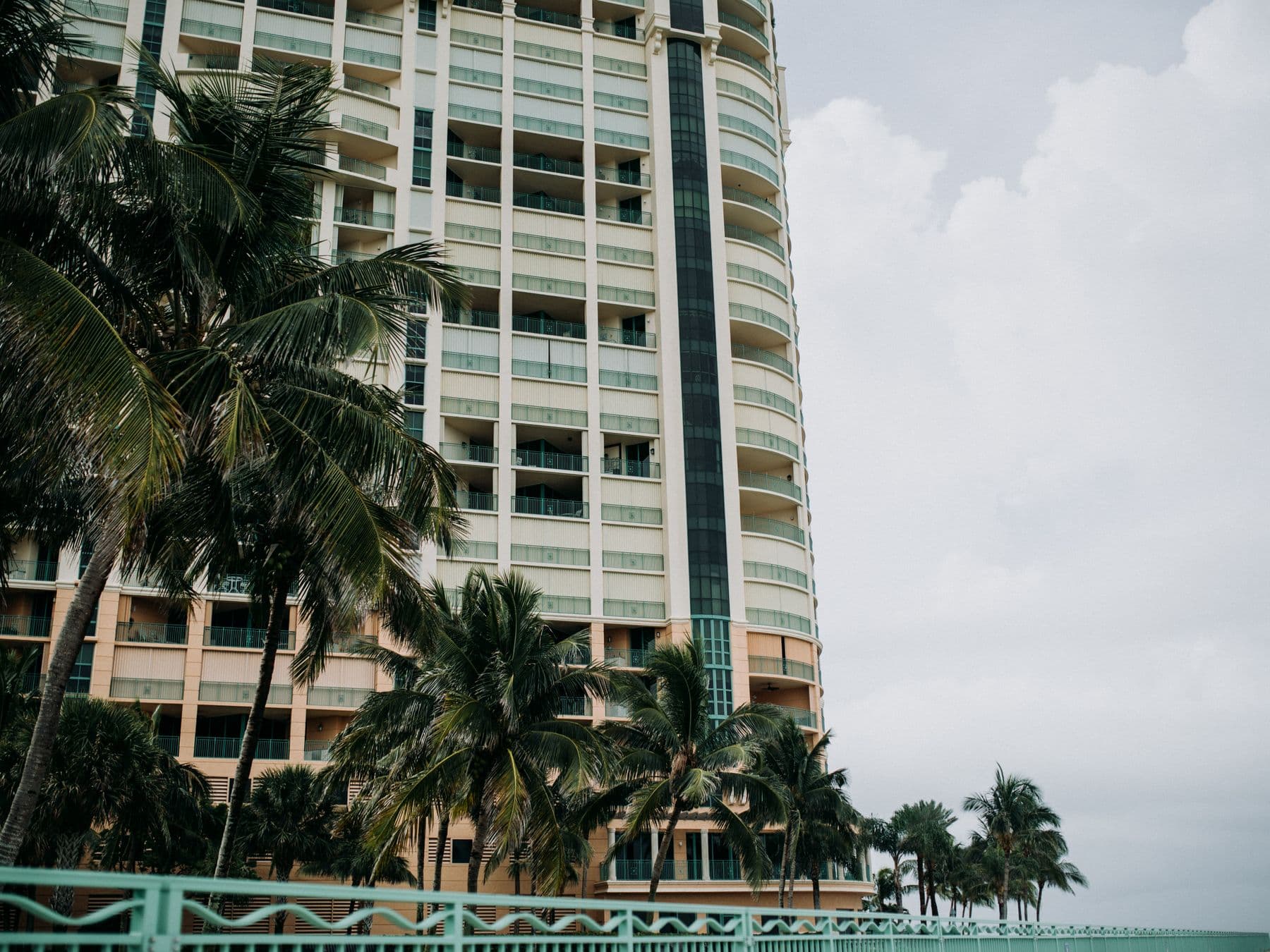 A tall residential tower with balconies behind palm trees and a waterfront railing.