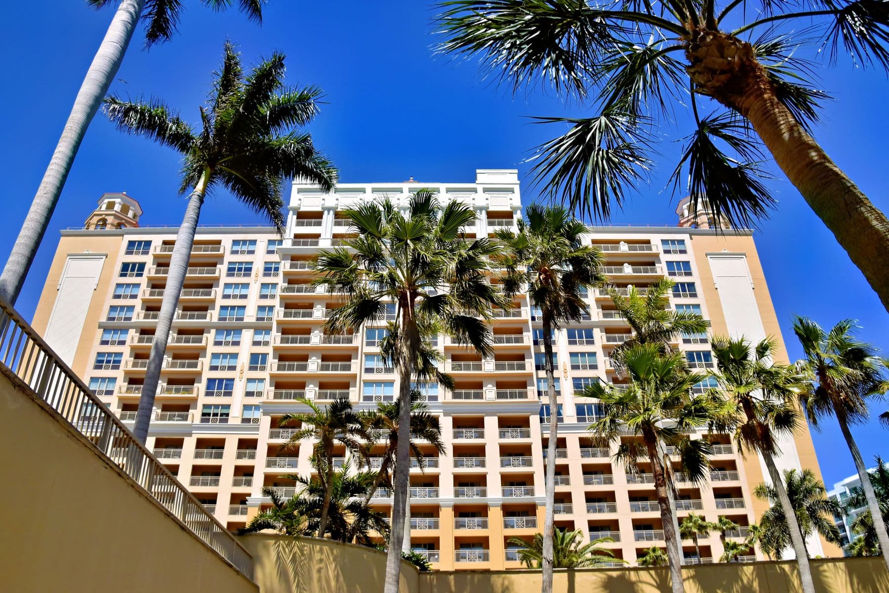 A tall cream and orange hotel with rows of balconies, seen from below between palm trees.