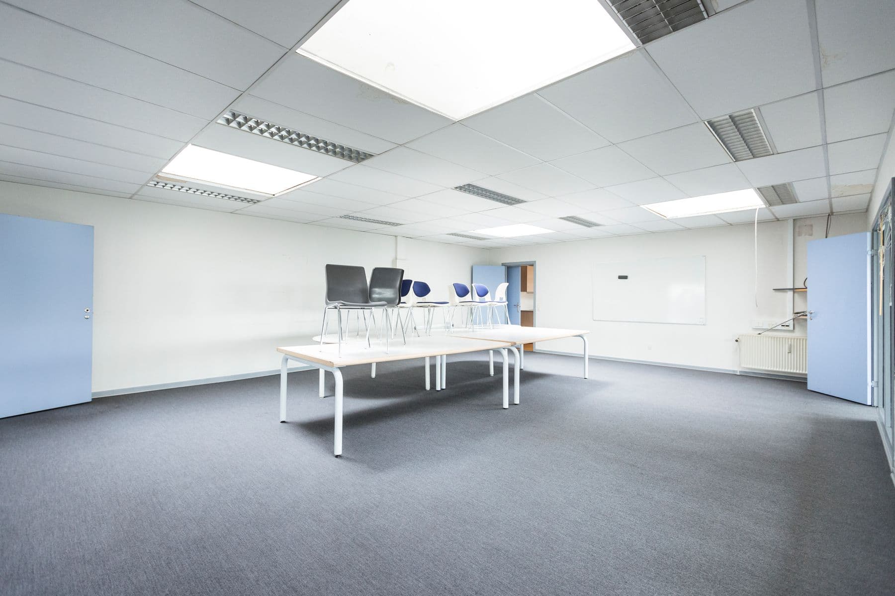 An empty office room with white walls, a gray carpet, ceiling lights and chairs stacked on tables.