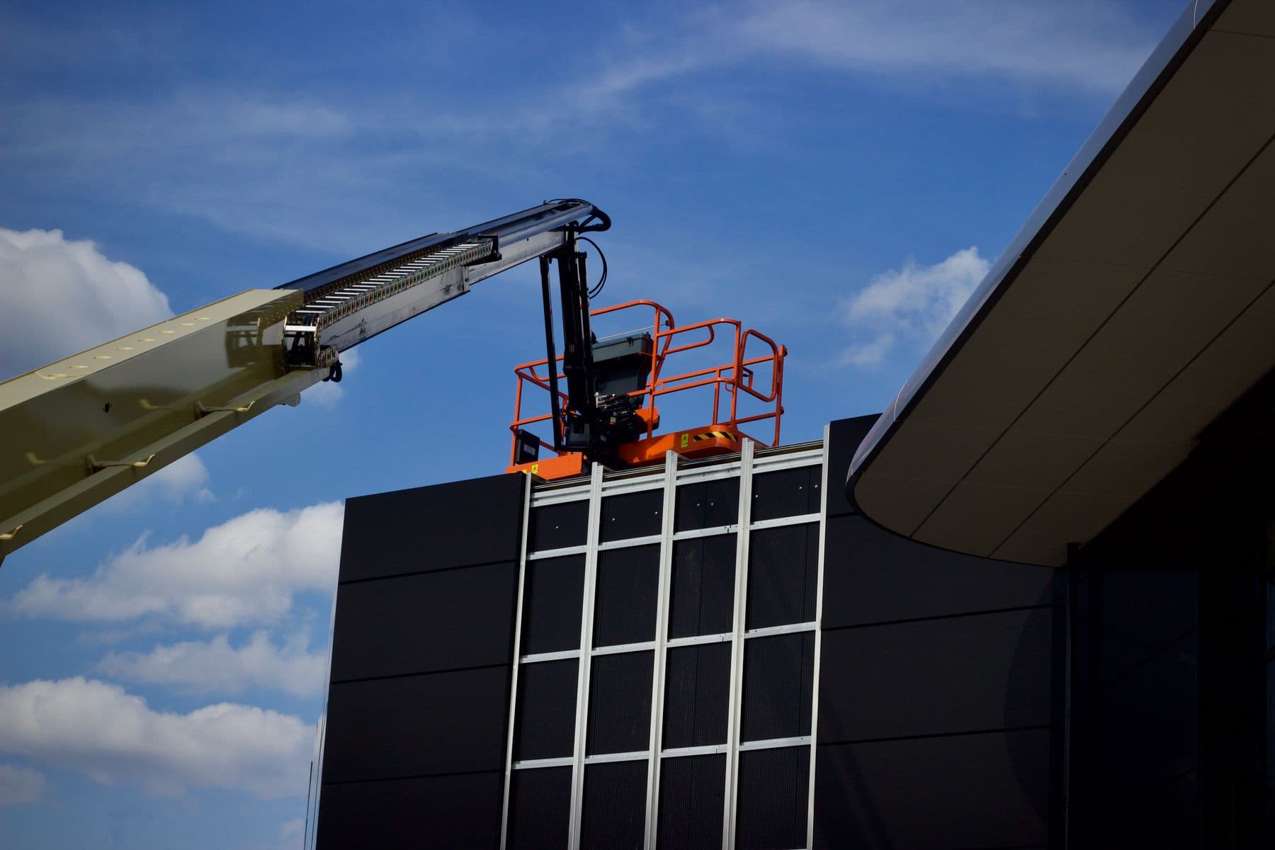 An orange boom lift basket raised above the dark glass facade of a modern building, under a blue sky.
