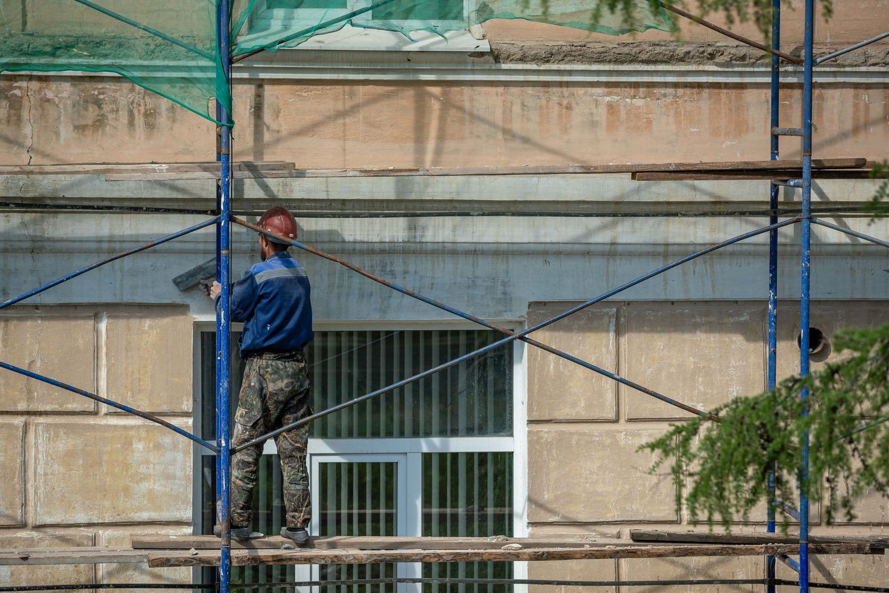 A worker in a hard hat on a blue scaffold, repairing the wall of an older building.