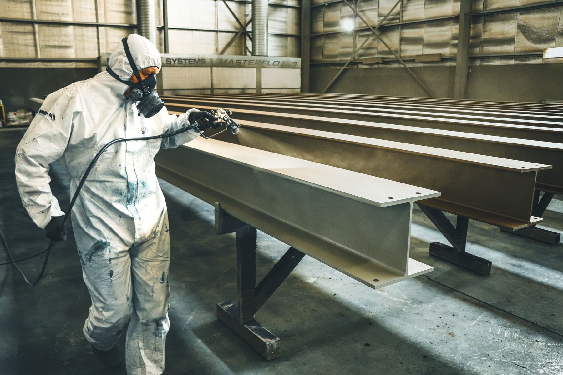 A worker in a white protective suit and respirator spraying coating on long steel beams indoors.